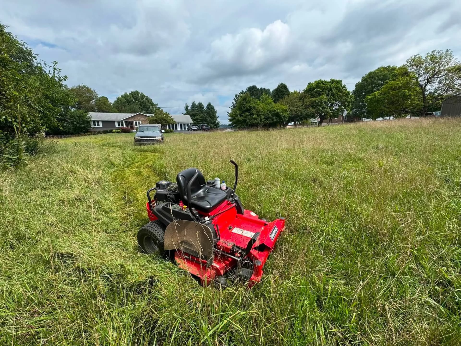 Red zero-turn mower cutting a path through tall grass in an overgrown field.